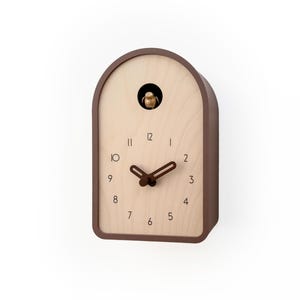Könnte beinhalten: Eine braune und beige Kuckucksuhr mit einem Vogel, der aus einer kleinen Öffnung kommt. Das Zifferblatt hat schwarze Ziffern und braune Zeiger. Die Uhr hat eine abgerundete Oberseite und einen rechteckigen Körper.