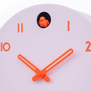 May include: A modern, pastel pink cuckoo clock with orange accents. The clock features orange numerals, hands, and a small bird inside a circular opening. The clock's design is minimalist and contemporary.