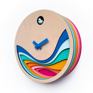 K&ouml;nnte beinhalten: Eine farbenfrohe Holzuhr mit geschichteten, wellenf&ouml;rmigen Ausschnitten in Blaut&ouml;nen, Gr&uuml;n, Gelb, Rosa und Orange. Das Zifferblatt der Uhr ist aus Naturholz mit blauen Zeigern.