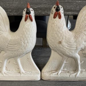 Puede incluir: Dos figuras de gallo de cerámica blanquecina. Cada gallo tiene una cresta y barbillas rojas, ojos negros y una cola detallada. Las figuras están sobre una base rectangular con un diseño texturizado. Estos gallos decorativos son adecuados para la decoración del hogar.