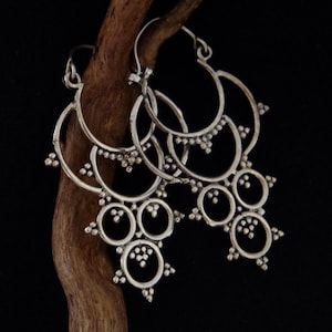 Könnte beinhalten: Silberfarbene Chandelier-Ohrringe mit mehreren kreisförmigen und halbkreisförmigen Elementen, verziert mit kleinen, perlenbesetzten Akzenten. Die Ohrringe werden auf einem dunklen Holzast vor schwarzem Hintergrund präsentiert, um ihr aufwendiges Design hervorzuheben.