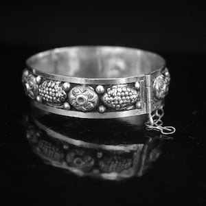 May include: A silver bracelet with a decorative design of repeating floral and geometric patterns. The bracelet has a hinged clasp.