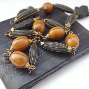 Può includere: Una collana vintage con perline color ambra e dettagli neri a coste. La collana è esposta su una superficie scura e strutturata, che mette in risalto il suo design intricato e l'estetica antica. La collana presenta dettagli in metallo dorato.