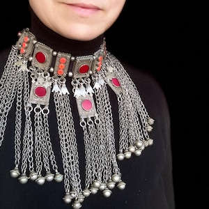 Può includere: Collana vistosa color argento con accenti rossi e corallo. La collana presenta pendenti quadrati e rettangolari con pietre rosse, collegati da catene a cascata. Piccole campanelle argentate pendono dalle catene, creando un effetto a strati.