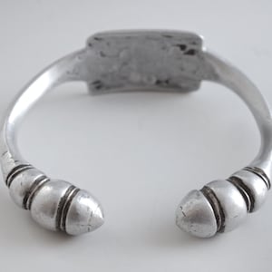 May include: Silver bracelet with a rectangular plate and two rounded ends with decorative ridges.