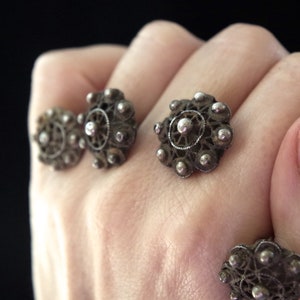 Puede incluir: Primer plano de pendientes antiguos de filigrana de plata. Los pendientes tienen un diseño floral con una cuenta central y cuentas más pequeñas circundantes. El intrincado trabajo de metal es visible sobre un fondo oscuro.