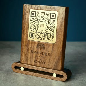 May include: A wooden QR code stand featuring a gold QR code and the text "RAFFLES LONDON THE OWO". The stand is made of dark wood and has a curved base with two gold-coloured accents.
