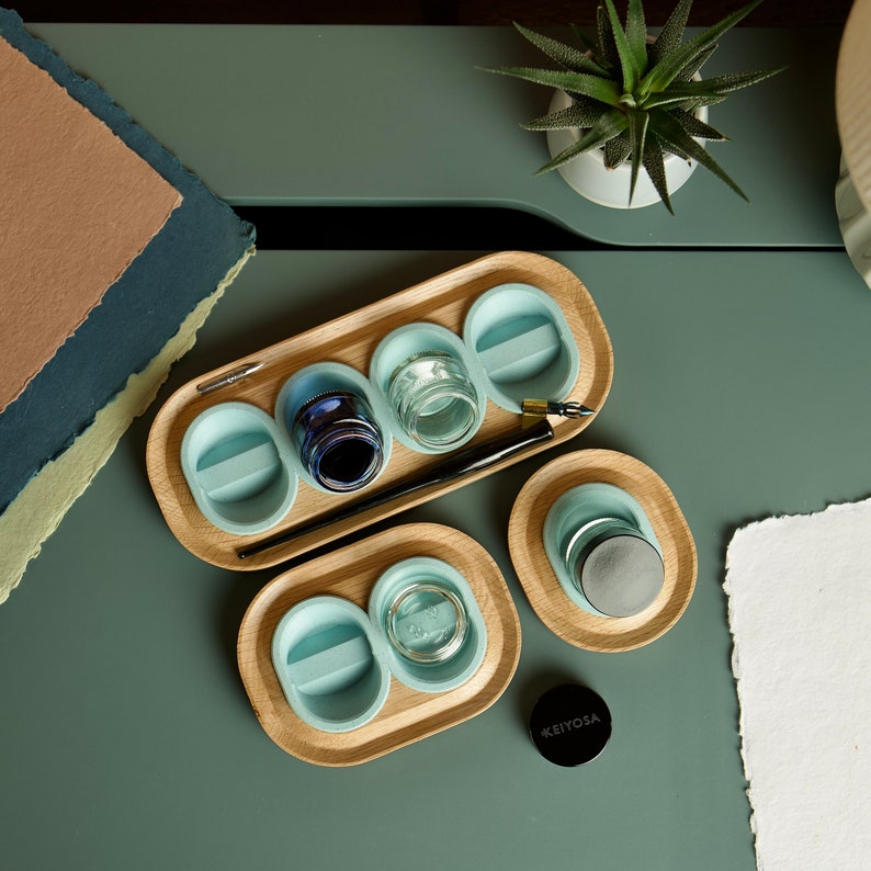May include: A wooden tray with six light blue wells for holding ink and other supplies. The tray is made of natural wood and has a light brown finish. The wells are arranged in a circle and are separated by a thin wooden divider. The tray is perfect for storing and organizing your calligraphy supplies.