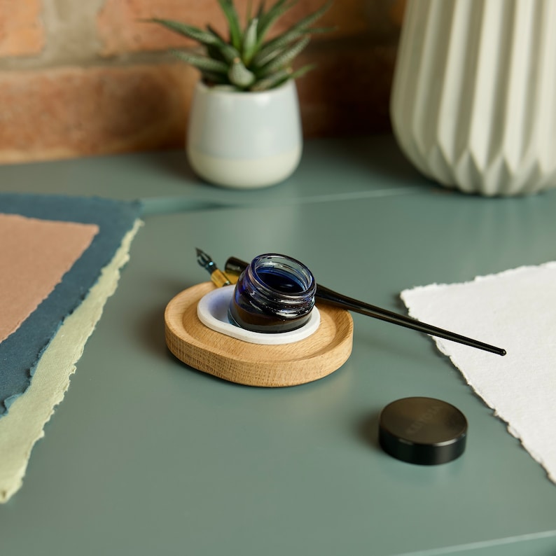 May include: A wooden inkwell holder with a blue glass inkwell and a black calligraphy pen. The inkwell is filled with dark blue ink. The holder is on a green surface with a piece of white paper and a stack of paper in the background.