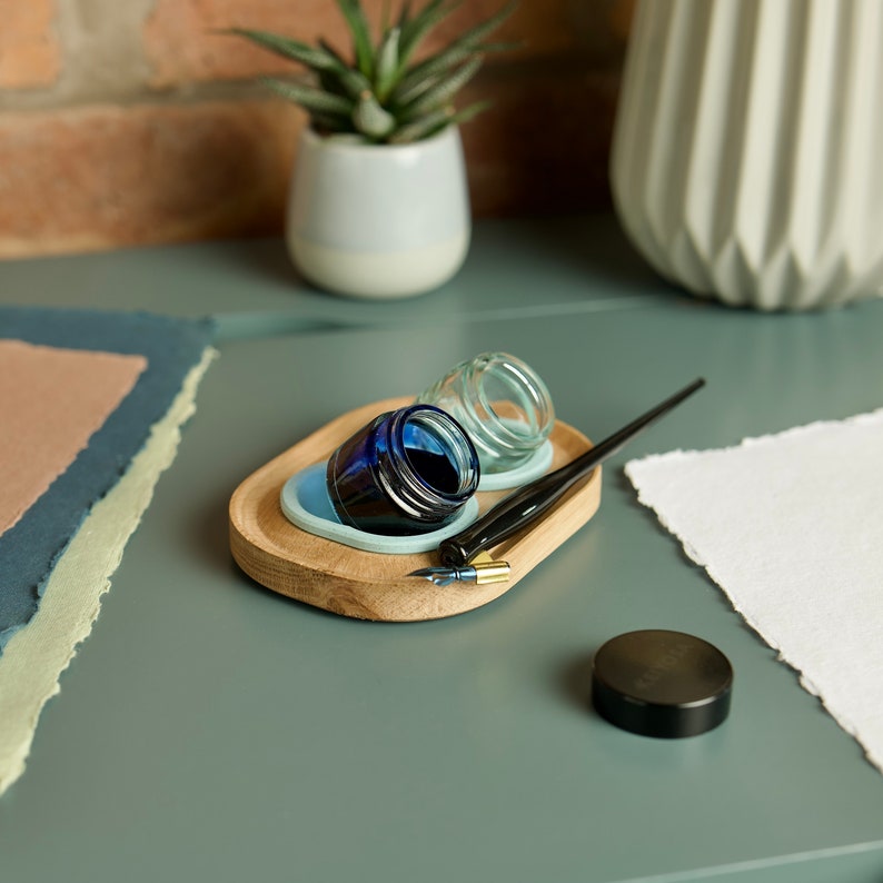 May include: A wooden tray with two small glass inkwells, one filled with blue ink, and a black calligraphy pen. The tray is on a light blue surface with a sheet of white paper.