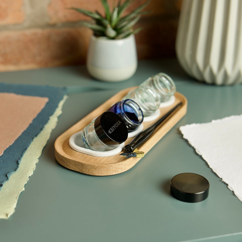 May include: A wooden tray with three glass inkwells and a black inkwell with the text "#KEIYOSA" on the lid. A calligraphy pen is on the tray. The tray is on a blue surface.