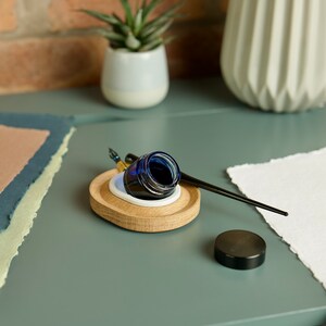 May include: A wooden inkwell with a blue glass ink bottle and a black calligraphy pen. The inkwell has a white ceramic insert and sits on a green surface.