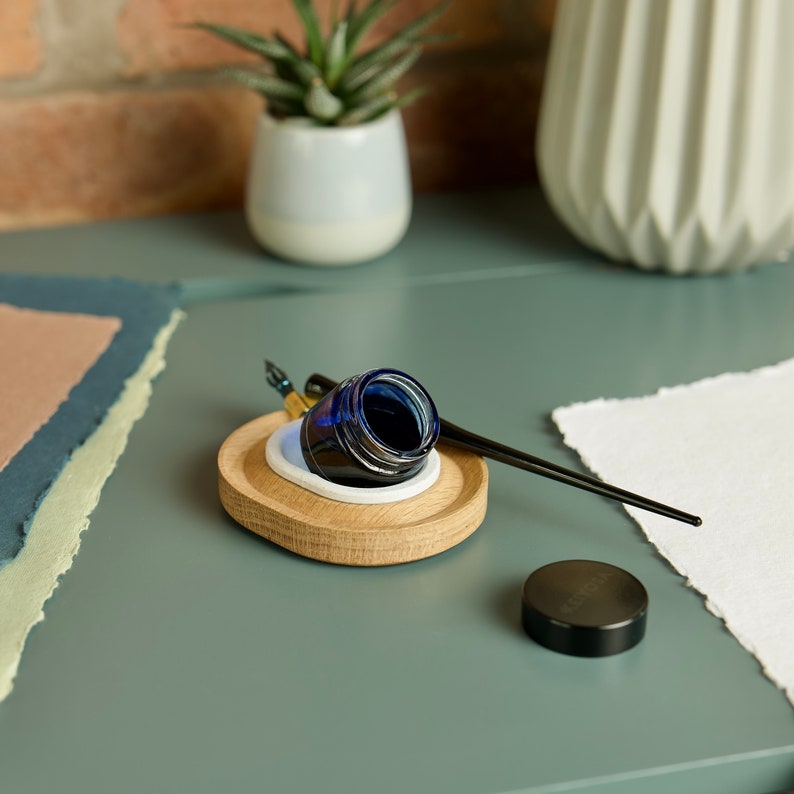 May include: A wooden inkwell with a blue glass ink bottle and a black calligraphy pen. The inkwell has a white ceramic insert and sits on a green surface.