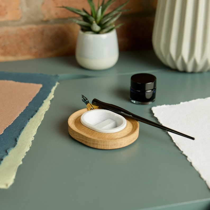 May include: A wooden calligraphy pen holder with a white ceramic well for ink. A black calligraphy pen rests on the holder. A small glass jar of ink sits on a blue surface.