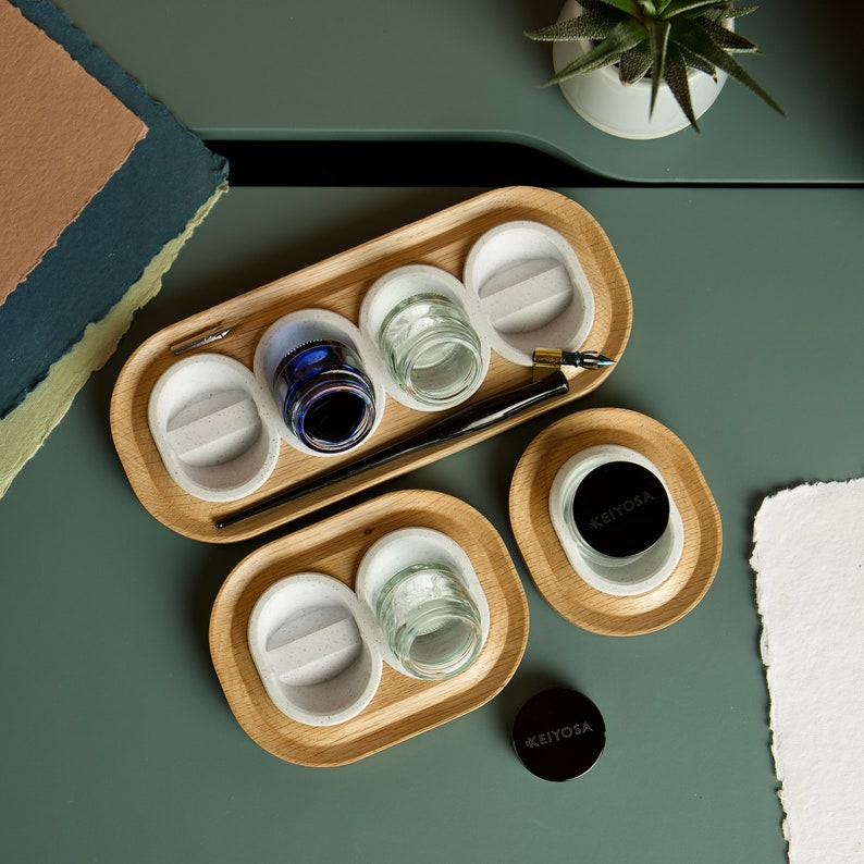 May include: A wooden tray with four small containers for ink or paint. The tray has a light wood finish and is divided into four sections. Each section has a small, round container with a white interior. The tray is perfect for storing and organizing small items.