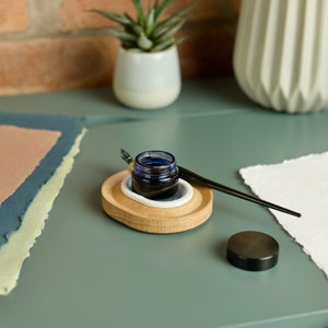 May include: A wooden inkwell holder with a white ceramic insert, a blue glass ink bottle, and a black calligraphy pen. The inkwell is on a teal surface with a stack of paper.