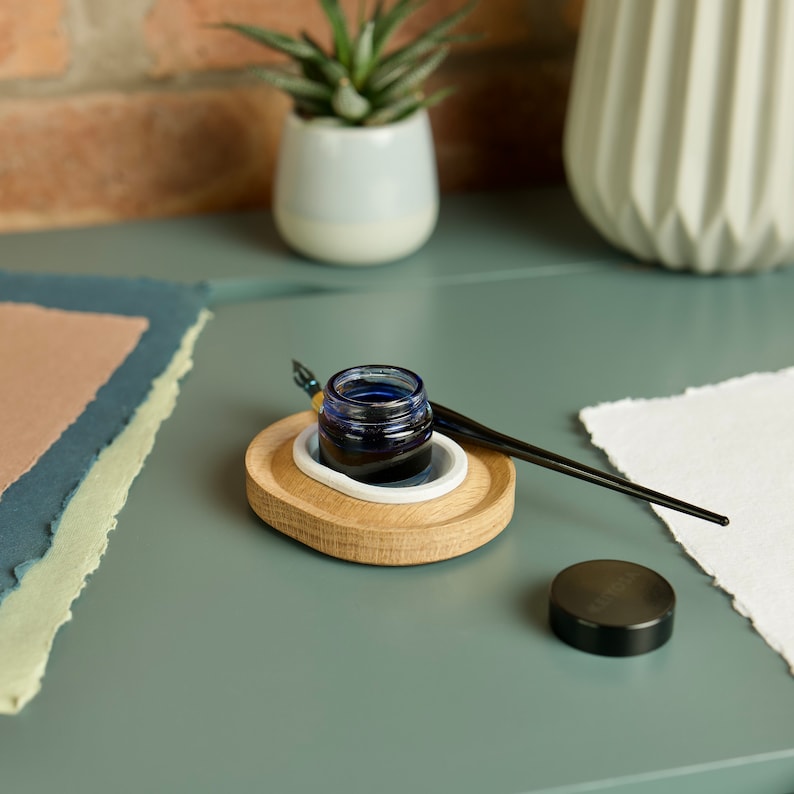 May include: A wooden inkwell holder with a white ceramic insert, a blue glass ink bottle, and a black calligraphy pen. The inkwell is on a teal surface with a stack of paper.