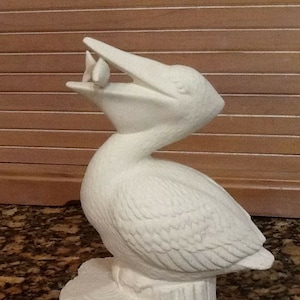 Op de afbeelding: Witte keramische pelikanfiguur met open bek, die een kleine vis in zijn bek houdt. De pelikan staat op een witte keramische boomstam.