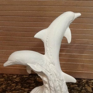 Puede incluir: Escultura de cerámica blanca de dos delfines saltando fuera del agua. Los delfines están colocados en una pose juguetona, con un delfín saltando sobre el otro. La escultura está detallada con olas texturizadas y una representación realista de los cuerpos de los delfines.