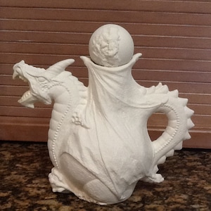 May include: White ceramic teapot shaped like a dragon with a round lid. The teapot is in the shape of a dragon with its wings spread out. The dragon's head is facing forward with its mouth open. The teapot is sitting on a brown surface.