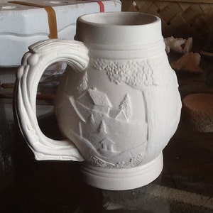 May include: White ceramic mug with a raised design of a winter scene with houses and trees. The mug has a large handle.