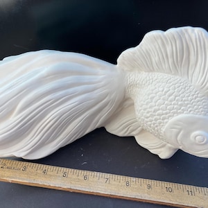 May include: A ceramic, unpainted goldfish figurine with detailed scales and flowing fins. The fish is lying on its side, showcasing its elaborate tail and dorsal fin. A ruler is placed near the fish for scale.