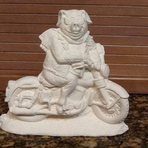 May include: White ceramic figurine of a pig wearing a leather jacket and riding a motorcycle. The pig is sitting on the motorcycle with its legs outstretched. The motorcycle has a large front wheel and a smaller back wheel.