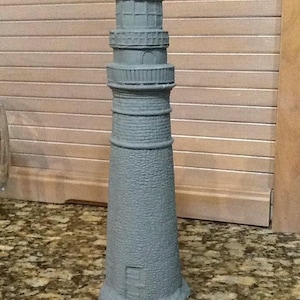 May include: A gray concrete lighthouse figurine with a detailed design. The lighthouse has a small window and a rounded top.