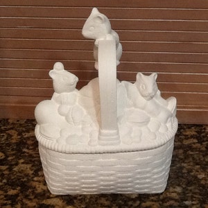 May include: White ceramic bisque basket with three small animal figurines. The basket has a woven design and a handle. The animals are sitting in the basket and appear to be squirrels.