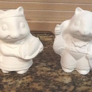 May include: Two white ceramic squirrel figurines, one carrying a fish and the other carrying corn. The squirrels are standing on a brown and gray countertop.