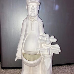 May include: A white ceramic figurine of a golfer wearing a cap and holding a golf bag with clubs. The figurine has a pocket in the front. The cap has a design on it.