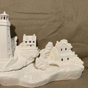 May include: A white ceramic coastal scene featuring a lighthouse, a house, and a building with a water wheel. The lighthouse is tall and cylindrical. The buildings are on rocky terrain, connected by a bridge. The scene is unpainted.