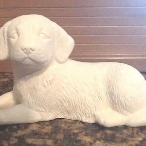 May include: A cream-colored ceramic puppy figurine in a reclining pose. The puppy has floppy ears, a detailed face, and textured fur. The figurine is resting on its front paws, with its body extended. The overall design is smooth and detailed.