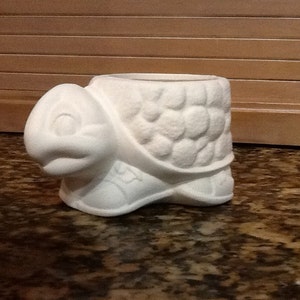 Puede incluir: Maceta de cerámica blanca en forma de tortuga con un diseño de caparazón texturizado. La tortuga está en posición de caminar con la cabeza girada hacia un lado.