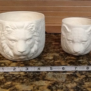 May include: Two unpainted ceramic planters. The larger planter features a detailed lion's face, while the smaller one has a cat's face. The larger planter is approximately 10 cm in diameter. The smaller planter is approximately 7.6 cm in diameter.