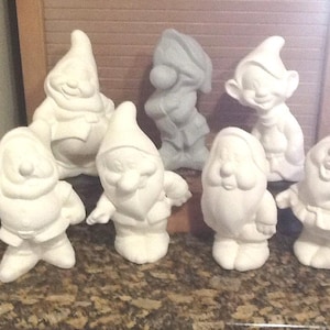 May include: Seven white ceramic figurines of dwarfs, each with a unique hat and facial expression. The dwarfs are standing on a brown surface.