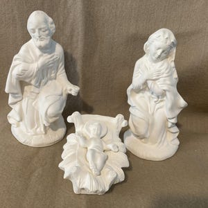 May include: A white ceramic nativity set featuring three figures: Joseph, Mary, and baby Jesus in a manger. The figures are kneeling or reclining, with detailed drapery and serene expressions. The set is designed for display.