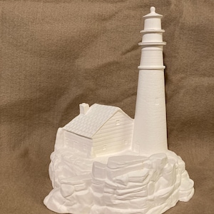 Puede incluir: Una figura de faro de cerámica blanca con una casa adjunta y una base rocosa. El faro tiene una torre alta y cilíndrica con bandas horizontales y una pequeña linterna. La casa presenta un tejado inclinado y revestimientos detallados.