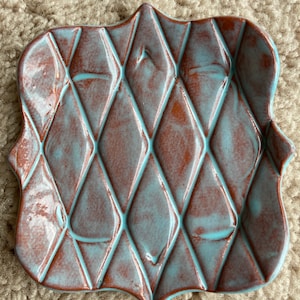 Puede incluir: Un plato de cerámica turquesa y marrón con un patrón de diamantes. El plato tiene un borde festoneado y es perfecto para servir aperitivos o guardar joyas.