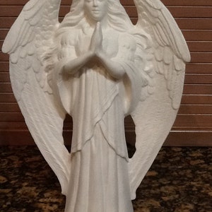Puede incluir: Figura de ángel de cerámica blanca con alas extendidas y manos juntas en posición de oración. El ángel lleva una túnica fluida y tiene el pelo largo y ondulado. La figura es un objeto decorativo.