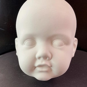 May include: White ceramic baby doll head with closed eyes, a small nose, and a slightly open mouth. The head is unpainted and ready for decorating.