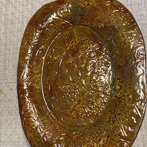 Puede incluir: Un plato de servir ovalado grande de cerámica con un esmalte marrón y verde. El plato tiene un diseño en relieve de intrincados patrones florales.