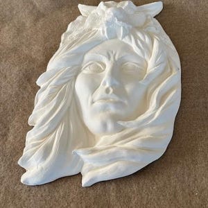 Peut inclure: Sculpture en relief blanche représentant un visage avec des cheveux fluides et une coiffe de loup. La sculpture présente des traits faciaux détaillés et est posée sur un fond neutre.