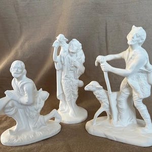 May include: Three white ceramic figurines depicting biblical figures. One kneels, another holds a lamb, and the third leans on a staff with a sheep. The figures are on individual bases, and the background is a neutral fabric.