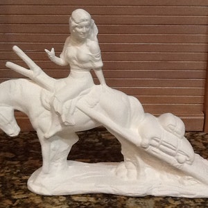 May include: A white ceramic sculpture of a woman riding a horse. The woman is wearing a dress and has long hair. The horse is rearing up, and the sculpture is on a base.