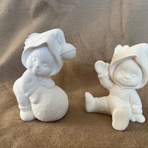 May include: Two unpainted ceramic figurines of children wearing hooded hats. One child is seated and holding a ball, the other is sitting with one arm raised. The figurines are white and appear ready for painting.