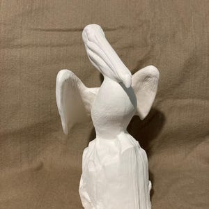 May include: A white ceramic pelican sculpture with outstretched wings. The bird is perched on a textured base, with its beak open. The sculpture is a solid white color and appears to be made of a plaster-like material. The pelican is a decorative item.