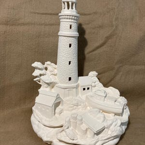 Puede incluir: Una escultura de faro de cerámica blanca con detalles intrincados. El faro presenta una torre alta, pequeños edificios y un barco, todo sobre una base rocosa. La escultura sin pintar muestra la textura y el diseño.