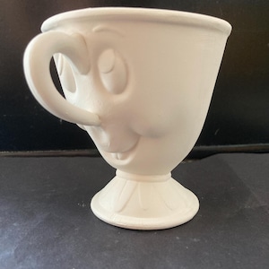 Könnte beinhalten: Eine weiße Keramik-Teetasse mit einem lächelnden Gesicht. Die Teetasse hat einen Henkel und steht auf einem Sockel. Die Teetasse ist unbemalt und kann individuell gestaltet werden. Die Teetasse ist etwa 10 cm hoch.
