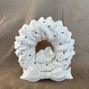 Könnte beinhalten: Ein weißer Keramik-Kranz mit einer Schleife unten und einem kleinen dekorativen Element in der Mitte. Der Kranz hat eine strukturierte Oberfläche mit kleinen schwarzen Punkten. Dieses Dekorationsobjekt eignet sich für die Inneneinrichtung.